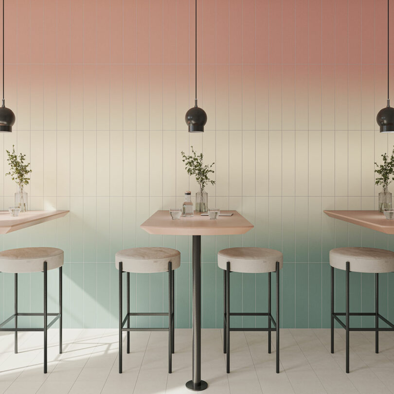 A minimalist café interior featuring tables with small potted plants, high stools, and gradient tiled walls transitioning from pink to cream to green, illuminated by black pendant lights.