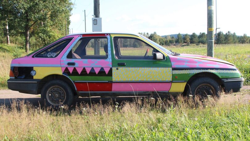 A side view of a car painted with a colorful, geometric pattern, parked on a grassy roadside with trees and a utility pole in the background