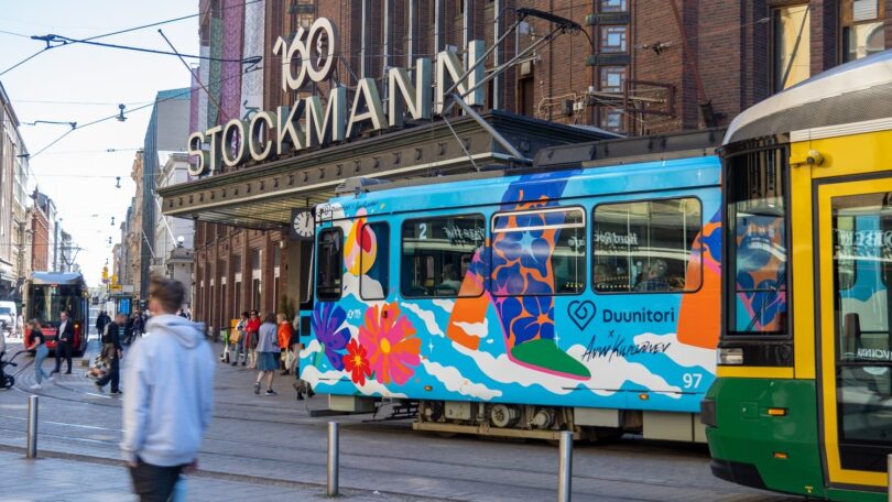 A colorful tram passes in front of the Stockmann department store. People walk on the street, and another tram is visible on the right