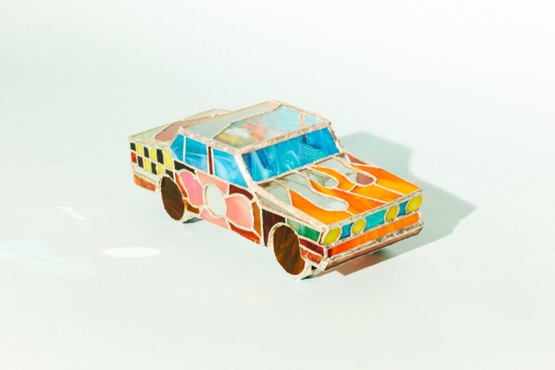 A colorful, abstract model of a car made from translucent materials, including various shapes of vibrant glass. The car features a boxy shape with highlighted front headlights