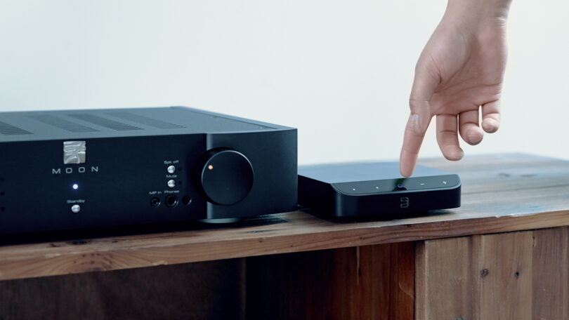 A person's finger is pressing a button on a small black electronic device beside a larger black audio amplifier, both placed on a wooden surface.