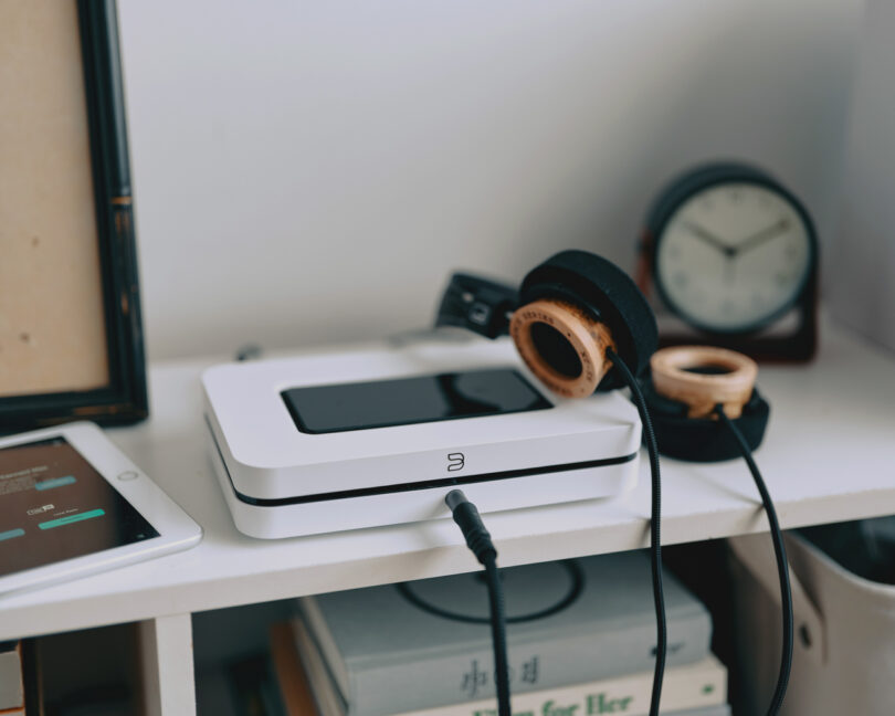 A pair of headphones rests on a white device with a display, connected by a black cable. Nearby are a tablet, a clock, and some books on a white shelf.