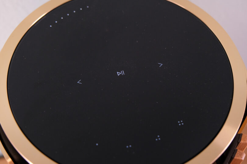 A close-up view of a round black touchpad with small white play, pause, and navigation symbols, encased in a gold-colored rim.