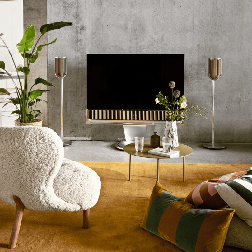 Modern living room with a large TV, stylish speakers, round coffee table with flowers and glasses, a cozy chair, and a green armchair on a mustard yellow rug. Plants and concrete wall in the background.