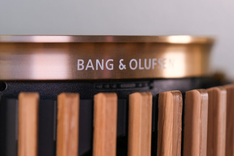 Close-up of a Bang & Olufsen logo on a product. The logo is etched on a circular metallic surface above a series of vertical slats.