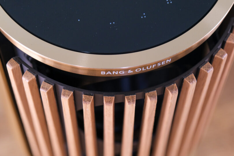 Close-up of a Bang & Olufsen speaker with a black top surface, surrounded by vertical wooden slats and a gold metal ring with the brand's name inscribed.