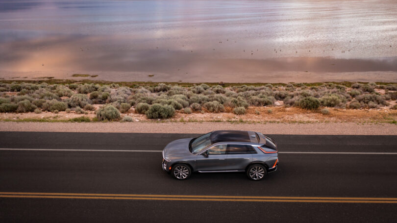 A gray SUV driving along a paved road next to a body of water with shrubs and sandy terrain on the roadside.