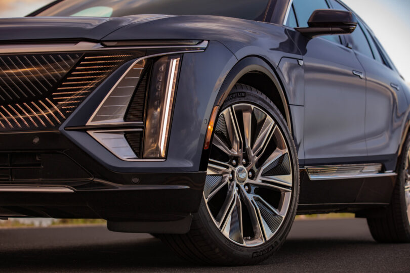 Close-up view of the front and side of a modern, dark-colored SUV, highlighting the sleek, angular headlight design, large alloy wheels, and sporty contours under a blue sky.