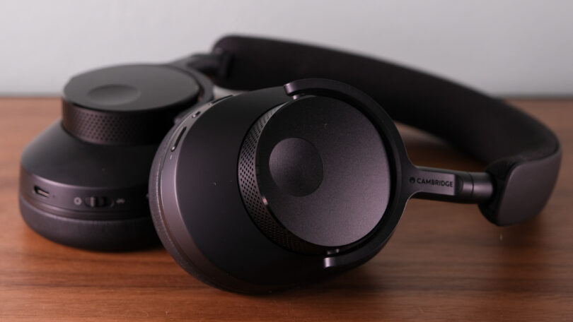 A pair of black over-ear headphones, known as Melomania P100, rests on a wooden surface. The brand name "CAMBRIDGE" is prominently displayed on one of the ear cups.