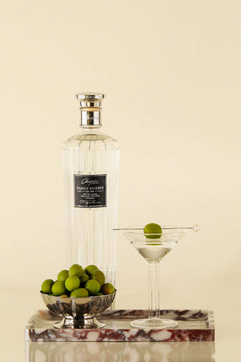 A clear bottle of vodka, a martini glass with a green olive, and a silver bowl of green olives on a marble slab