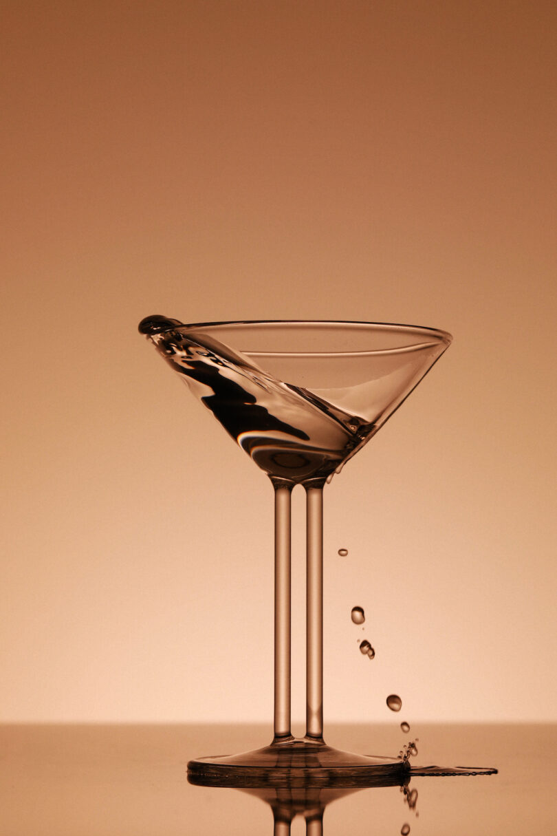 A martini glass is tilted, causing liquid to pour out and create droplets midair against a beige background