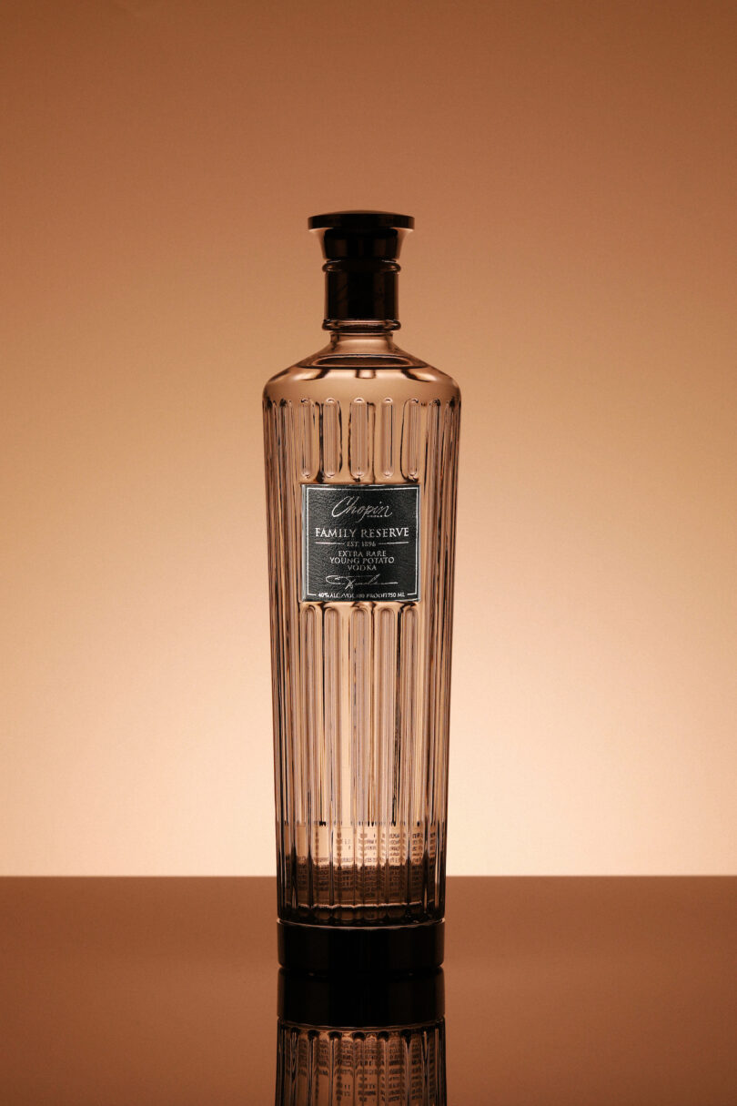 A clear, decorative glass bottle with a black label and cap, set against a warm gradient background