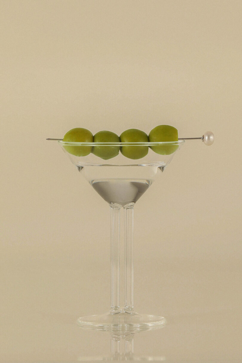 Clear martini glass with four green olives skewered on a pick, sitting on an off-white background