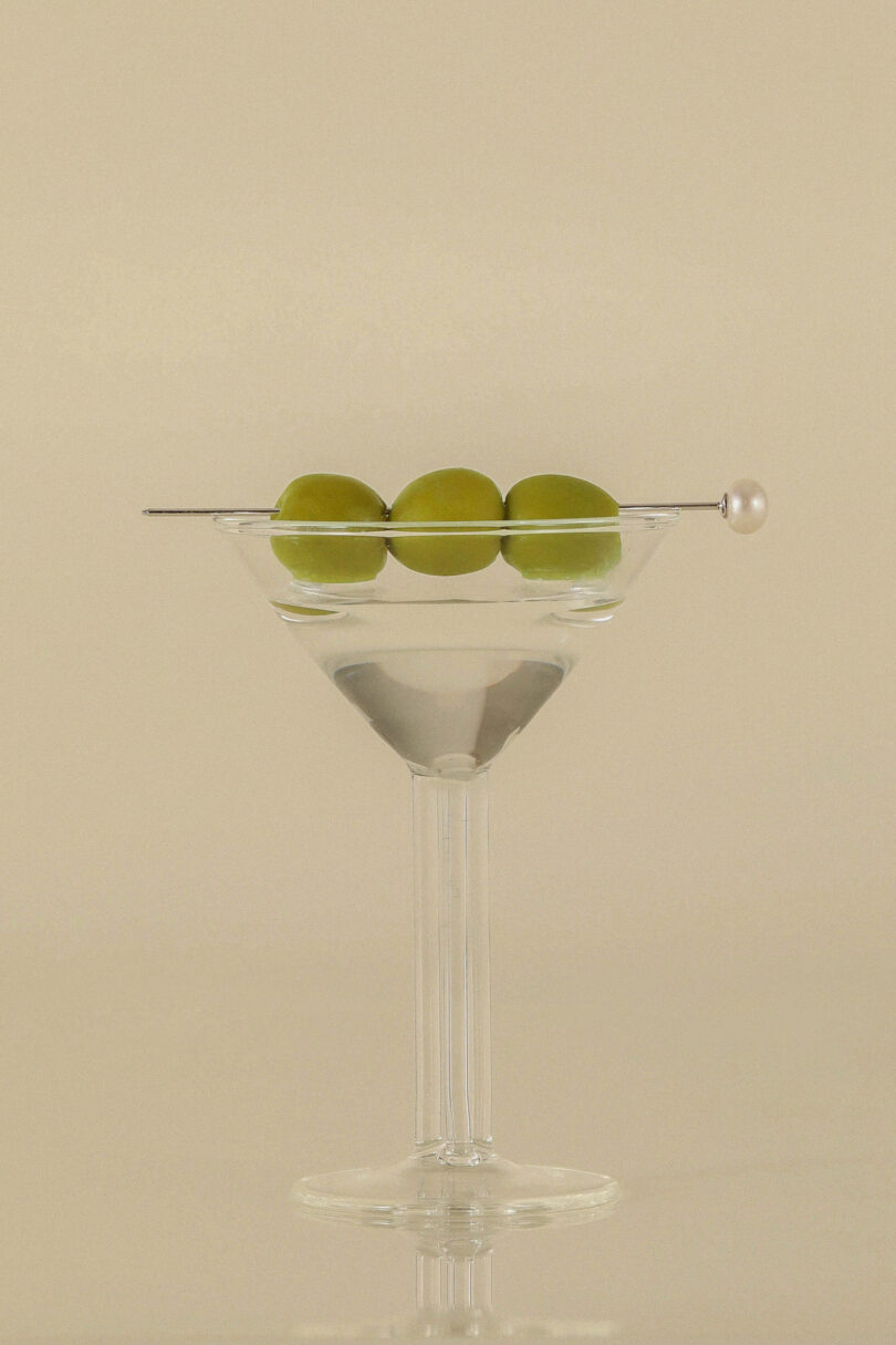 Clear martini glass with three green olives skewered on a pick, sitting on an off-white background