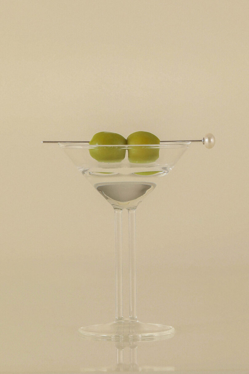 Clear martini glass with two green olives skewered on a pick, sitting on an off-white background