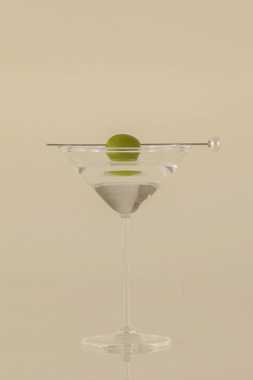 Clear martini glass with a single green olive skewered on a pick, sitting on an off-white background