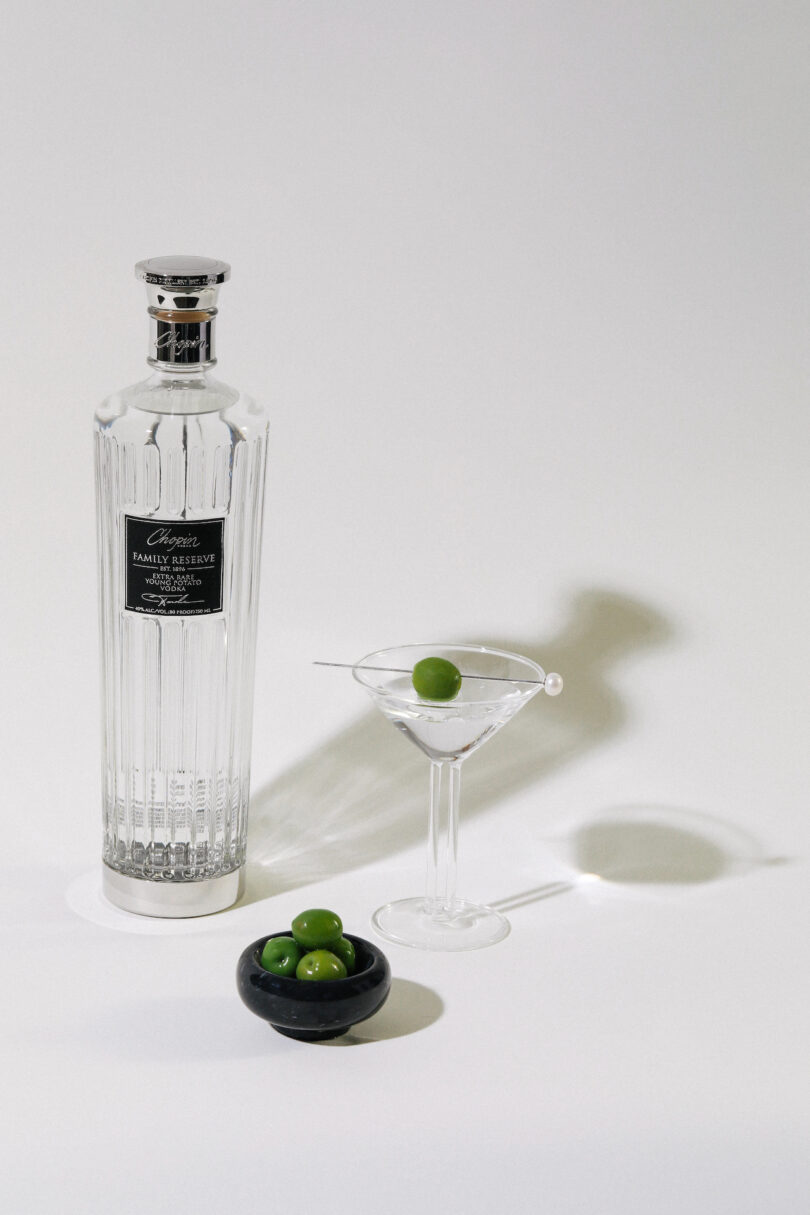 A bottle of gin, a martini glass with an olive garnish, and a small bowl containing four green olives on a white surface