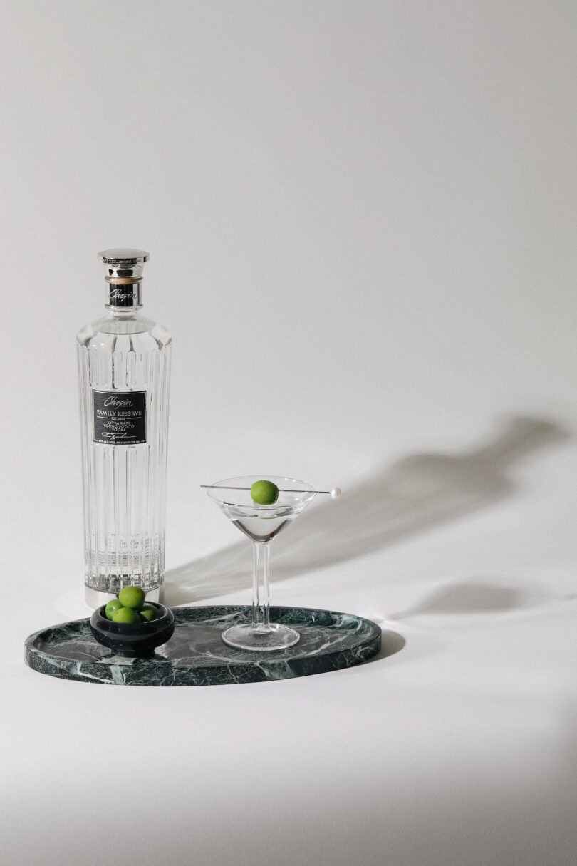 A bottle of gin, a filled martini glass with an olive garnish, and a small bowl with two olives are set on a green marble tray