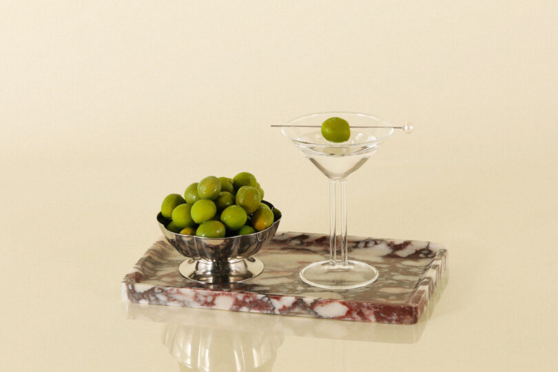 A silver bowl filled with green olives and a martini glass with an olive on a skewer are placed on a marble slab