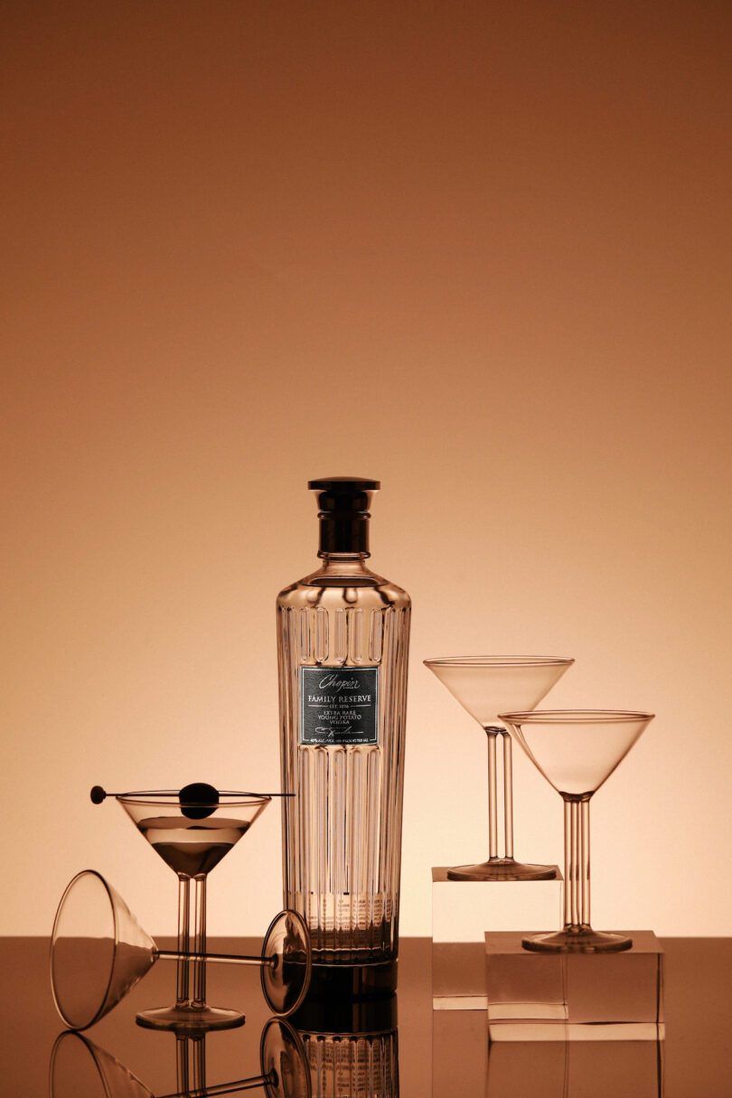 A tall, clear-cut glass bottle with a black cap stands beside two empty martini glasses and one filled martini glass with an olive garnish against a warm, gradient background