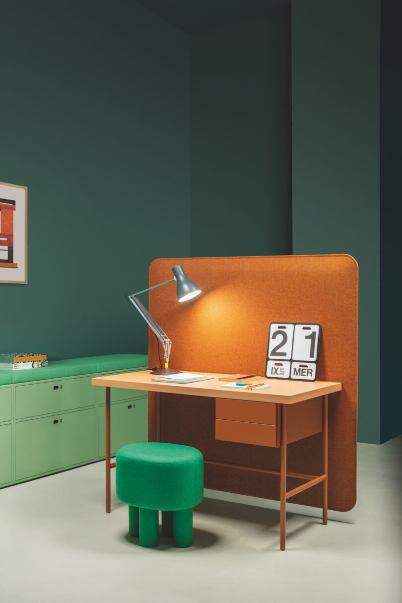 A minimalist office setup with a wooden desk, green stool, desk lamp, and a flip calendar displaying the 21st. The backdrop is a mix of green cabinets and a dark teal wall