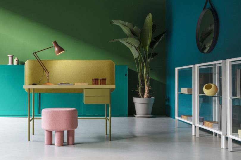 A modern office space features a green desk with a pink stool, a potted plant, and a wall-mounted mirror. A bookshelf with various items is situated on the right