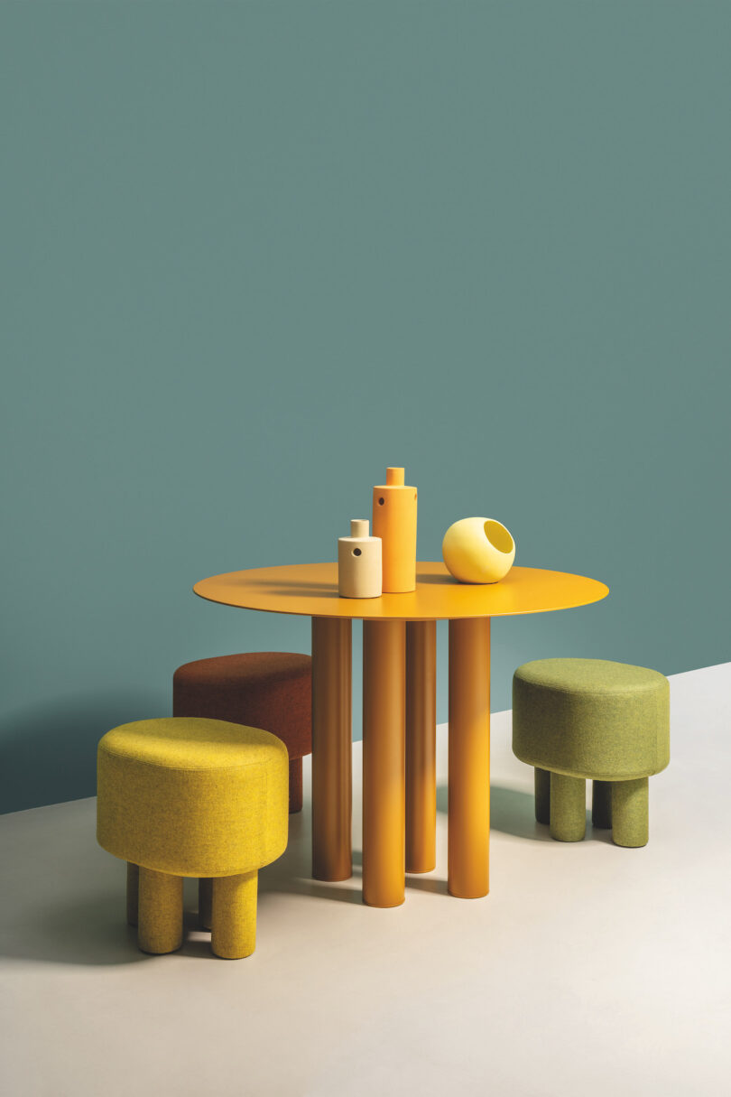 A round yellow table with three yellow, green, and orange stools are arranged against a teal wall. Various yellow and beige vases are on the table