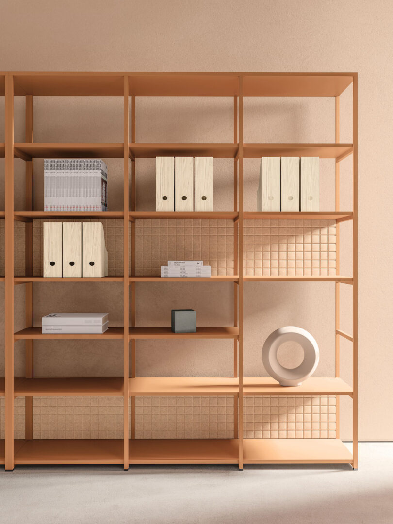 A minimalist wooden bookshelf against a beige wall, holding a few ring binders, books, a small box, and a circular sculpture