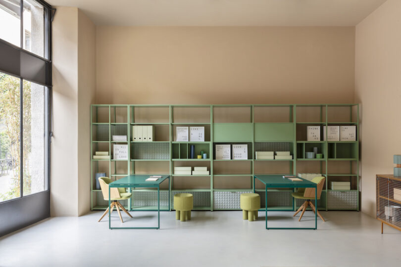 A modern office with green shelves, two desks, and chairs. The shelves hold books, folders, and decorations. Large windows allow natural light into the room. Beige walls create a calm atmosphere