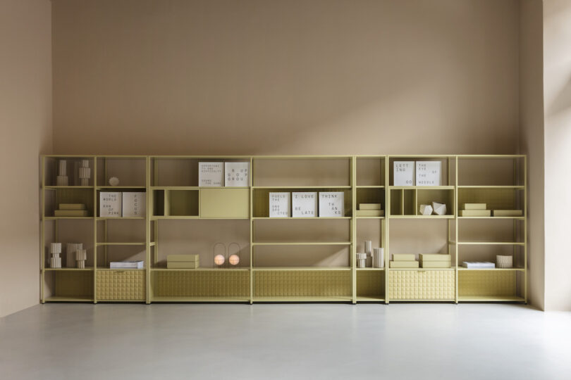 A beige-colored shelving unit against a neutral wall holds various minimalistic decorative items including boxes, books, and small objects. The setting is well-lit with natural light