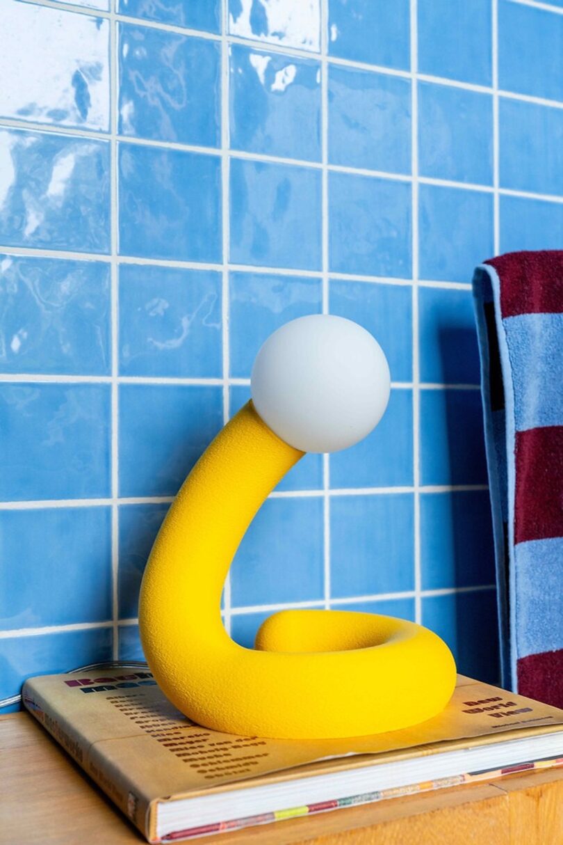 A yellow, snake-like lamp with a white bulb on top, placed on a wooden surface against a blue tiled wall. A red and blue striped towel hangs nearby