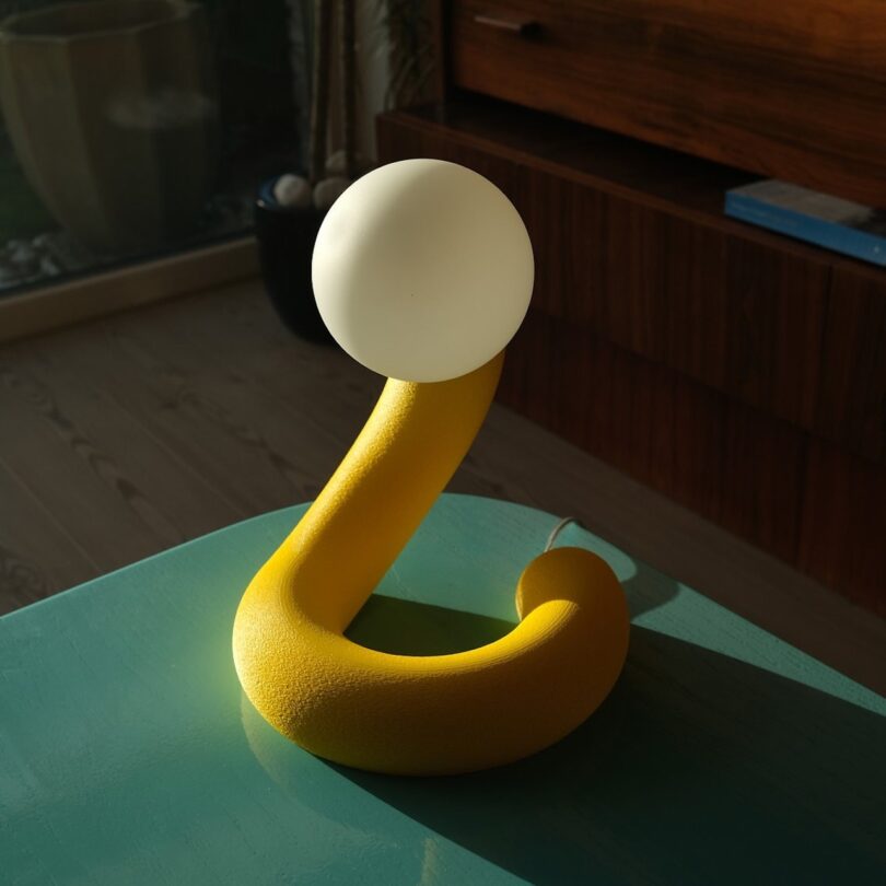 A yellow, curved table lamp with a spherical white shade sits on a teal surface in a room with wooden furniture and a glass window