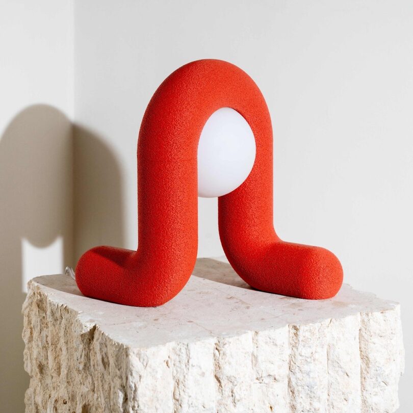 A modern, red sculptural lamp with a unique U-shaped design, resting on a textured stone pedestal. A white spherical light bulb is at the center of the U-shape