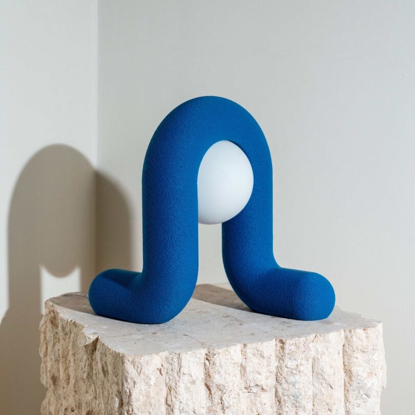 A modern sculpture featuring a U-shaped, blue textured structure with a white spherical object in the center, displayed on a rough, beige stone pedestal