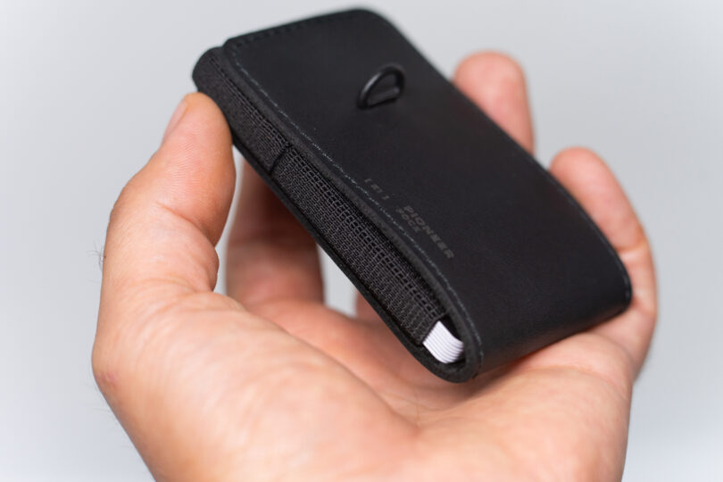 A hand holding a black leather wallet with a visible elastic band and a small metal loop on top.