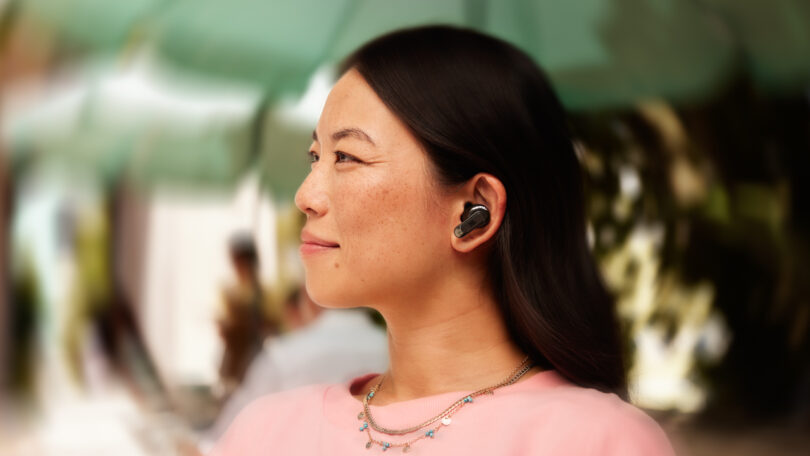 A woman with long dark hair is outside under an umbrella, wearing wireless earbuds. She is looking to the side and is dressed in a light pink top with a silver necklace.