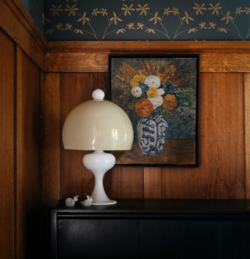 A mushroom-shaped white lamp on a black dresser next to a framed painting of a flower arrangement in a vase on a wooden paneled wall