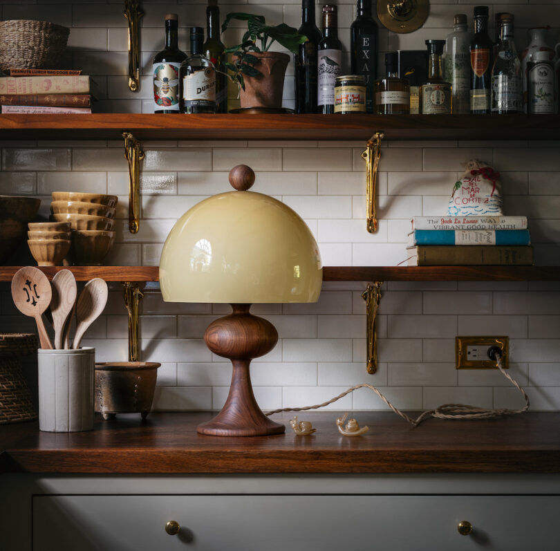 A kitchen counter with various objects including a wooden-based table lamp with a yellow dome shade, wooden utensils in a holder, and shelves with bottles, books, and plants against tiled walls