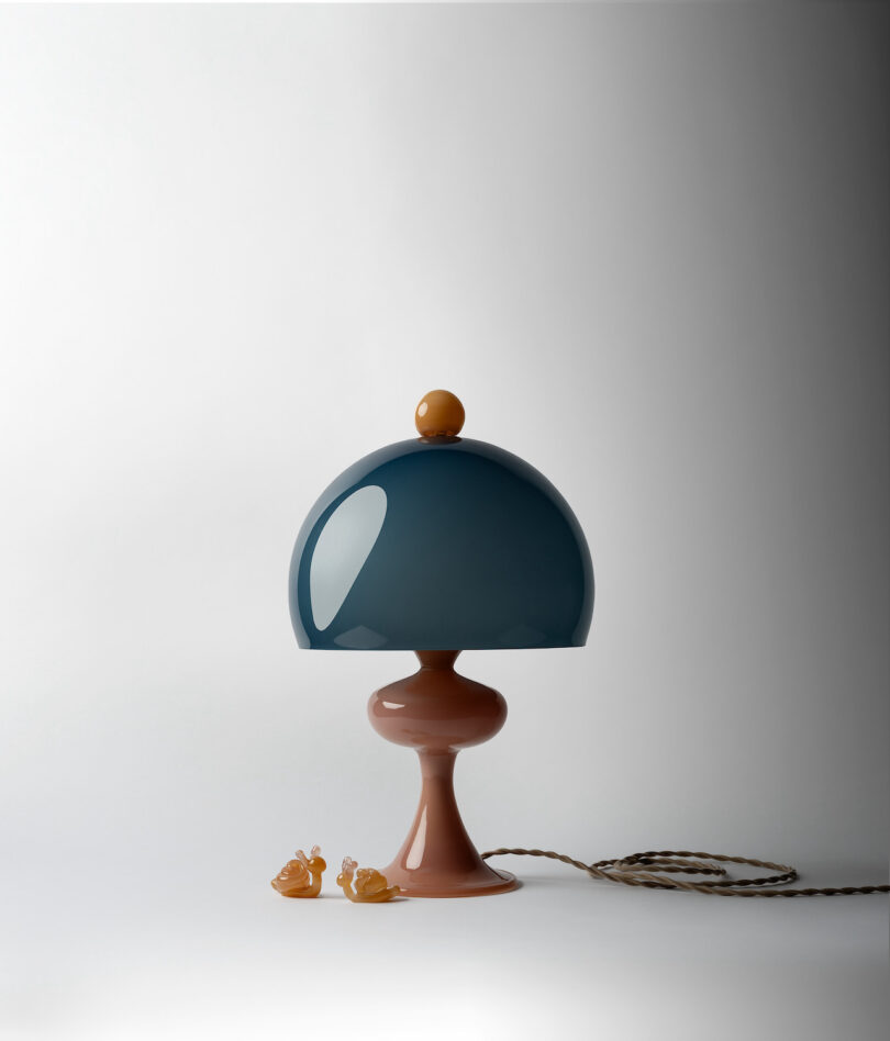 A blue dome-shaped lamp with a bulbous brown base and cord sits on a white surface with two small snail figurines at its base