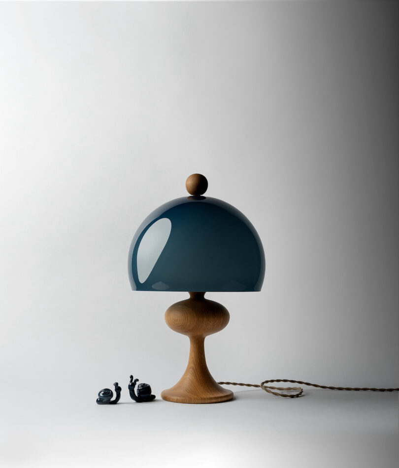 A wooden lamp with a dark green dome-shaped shade is placed beside two small figurines of snails on a clean, minimalist background