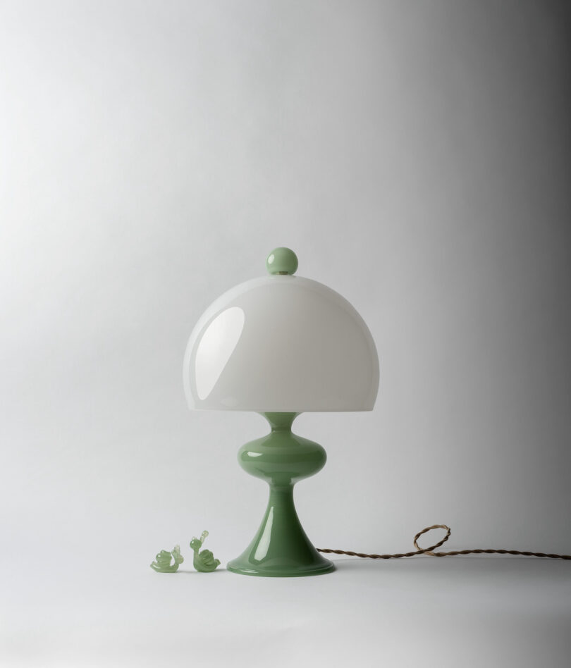 A green and white table lamp with a dome-shaped shade stands on a neutral background, accompanied by two small green glass snail figurines. A twisted cord is visible, trailing from the lamp.