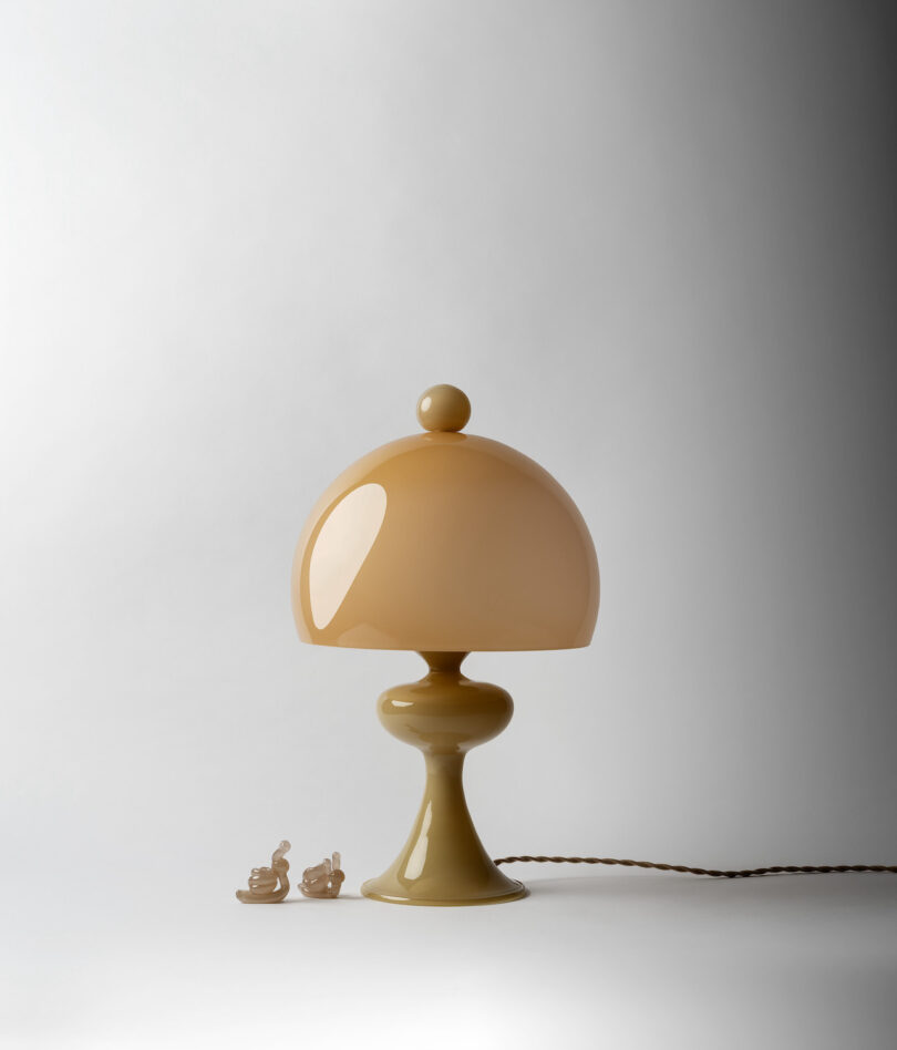 A beige, mushroom-shaped table lamp with a round finial on top is placed on a surface. There are two small ceramic snail figurines beside it.