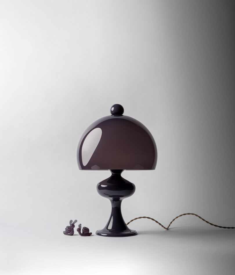 A modern black table lamp with a rounded shade and a thick base stands on a white surface. A twisted cord extends from the lamp, and two small snail figurines are placed nearby