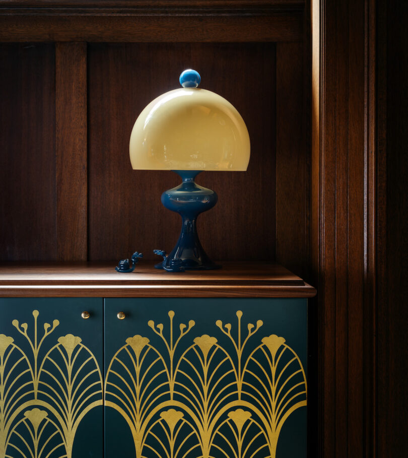 A table lamp with a round beige shade and dark blue base sits on a wooden cabinet adorned with a gold floral pattern