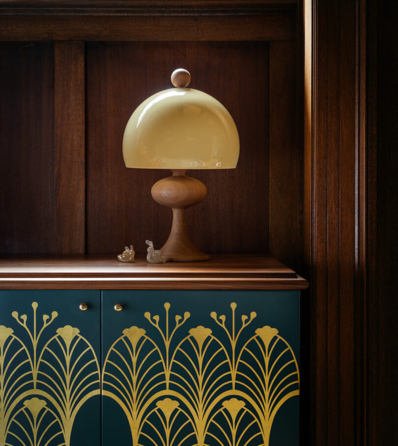 A wooden cabinet with a dark wooden frame features an art deco design with gold detailing. A modern beige table lamp with a wooden base sits on top, accompanied by small figurines