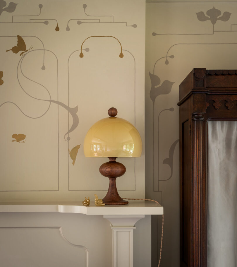 A yellow lamp on a white mantle shelf next to a brown wooden cabinet, with wall decor featuring butterflies and stylized leaves