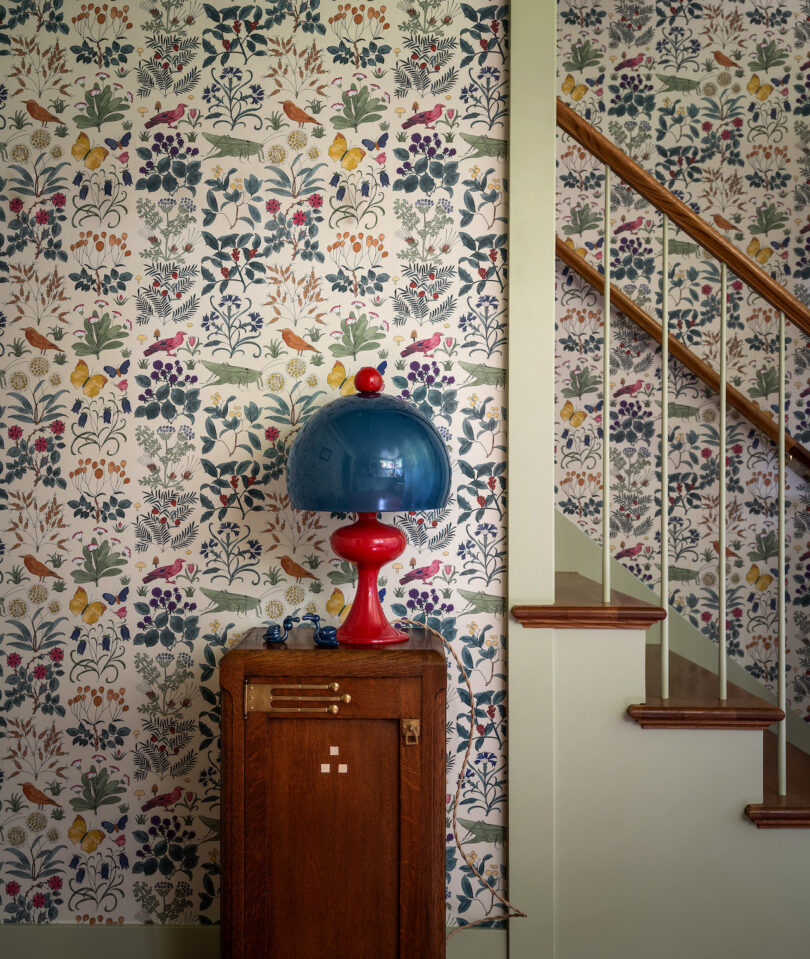 An interior scene with floral wallpaper, a wooden cabinet with a blue lamp on top, and a staircase with a wooden banister