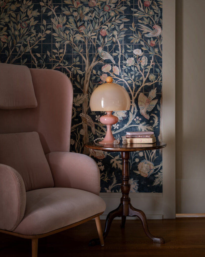 A cozy corner features a pink armchair next to a small wooden table with a pink lamp and books. The backdrop is a floral wallpaper with birds and branches