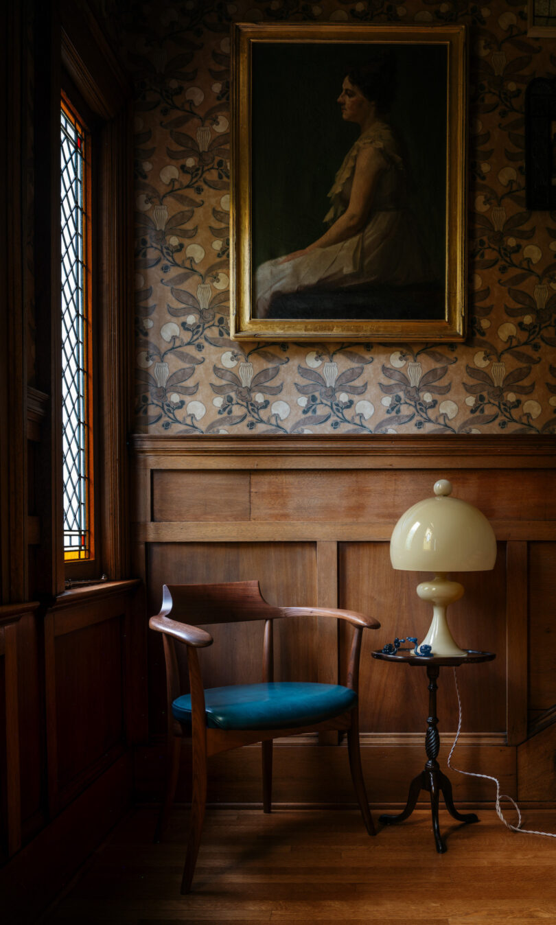 A corner of a room with wooden paneling includes a wooden chair with a blue cushion, a small lamp on a side table, and a portrait of a woman hanging on floral patterned wallpaper