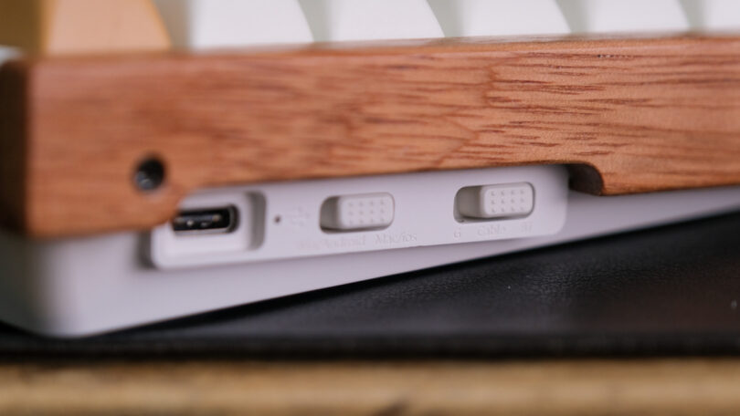 Close-up of the side of a keyboard showing a USB-C port and three switch options labeled "Mac/PC", "K6/G6", and "Cable/Bluetooth" set into a wooden frame.